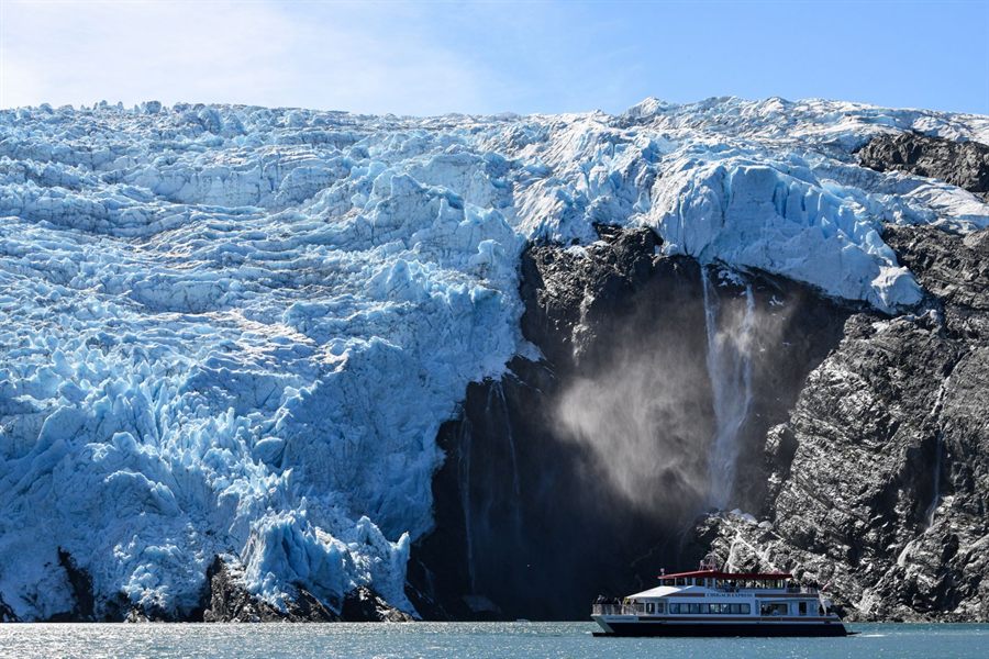 Waterfall pouring down beside a tidewater glacier in Alaska with a small boat below